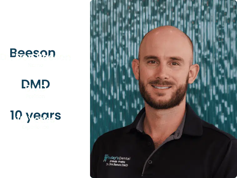meet-our-team-dr-don A man with a beard and a shaved head smiles at the camera, standing in front of a blue patterned background. He is wearing a black collared shirt with embroidery, sharing his warm smile at Dental Grande Prairie.
